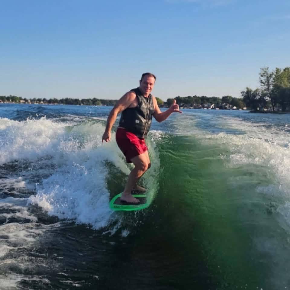 John Tremaine wakesurfing on a sunny day, giving a shaka sign